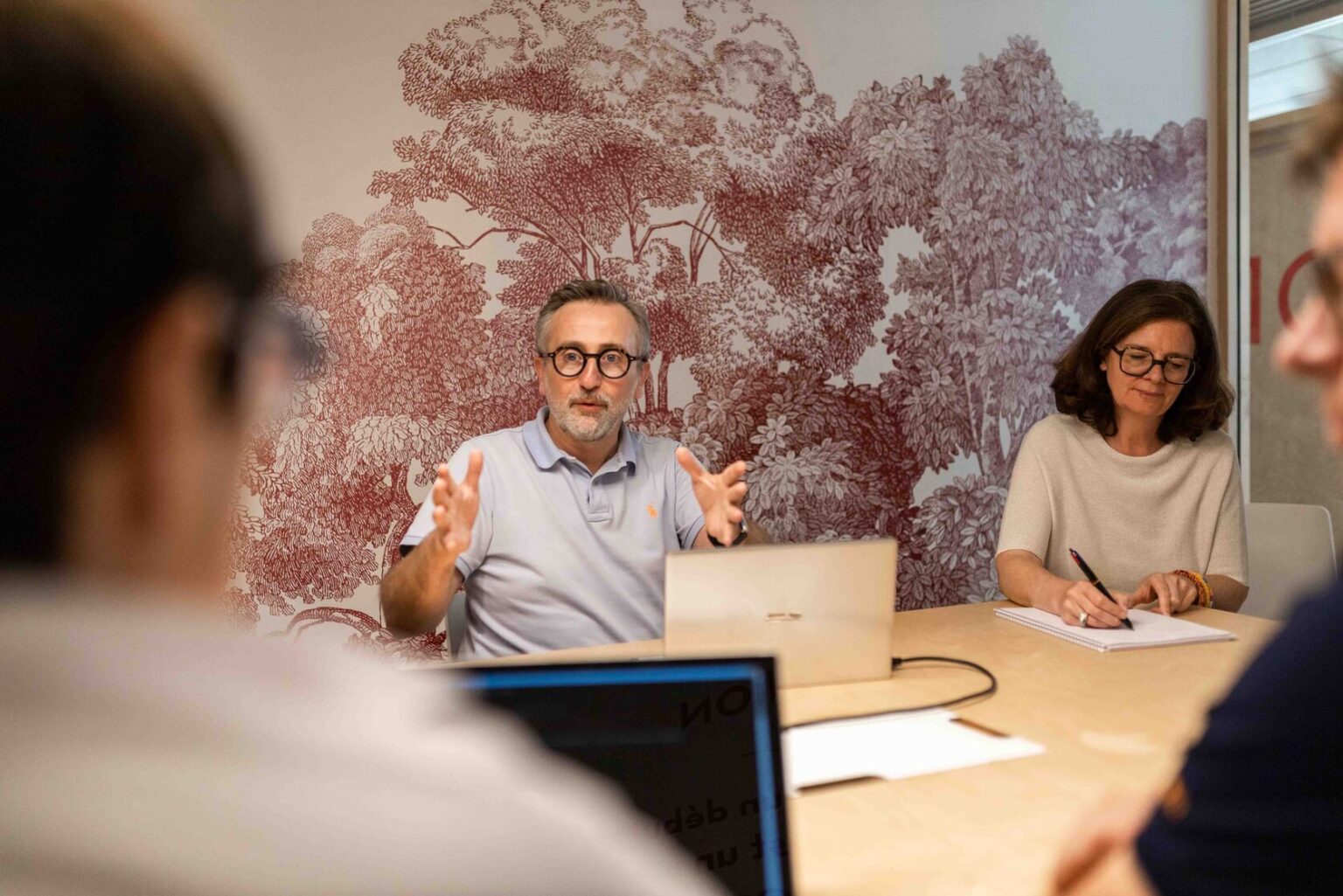 Un expert de chez Kubs expose et explique sa vision à deux clients assis face à lui. Impliqué et proche de ses clients il utilise ses mains pour appuyer ses conseils. A ses côtés, une employées de Kubs prend des notes pour son dossier de suivi