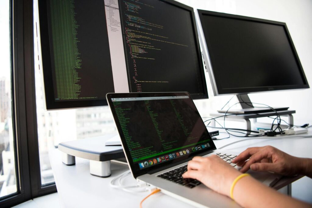 les mains d'une personne en train de taper sur le clavier d'un PC Portable allumé et, en surplomb, deux grands écrans allumés sur lesquels on dévine des graphiqiues et informations