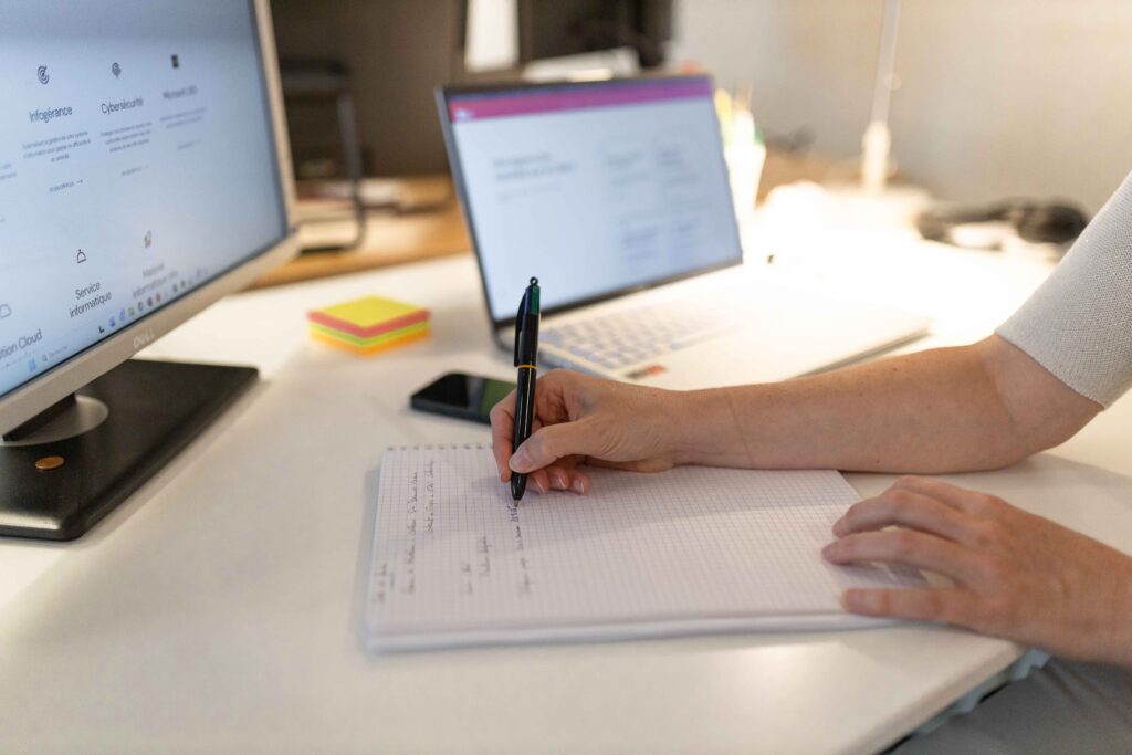 Gros plan sur la main d'une personne qui écrit sur un cahier posé sur un bureau. En arrière plan il y a 1 écran d'ordinateur allumé. Sur l'écran iplacé face à la personne qui écrit, on lit distintement Infogérance, Cybersécurité, Service informatique , cloud, Microsoft, matériel informatique.