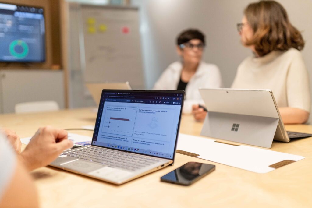 Au premier plan : un personne assise à une table de réunion devant son PC portable ouvert avec des graphiques sur l'écran. En face, uen employées de Kubs écoute une client qui lui parle. On devine au fond de la pièce, une présentation sur un grand écrand et un paperboard.