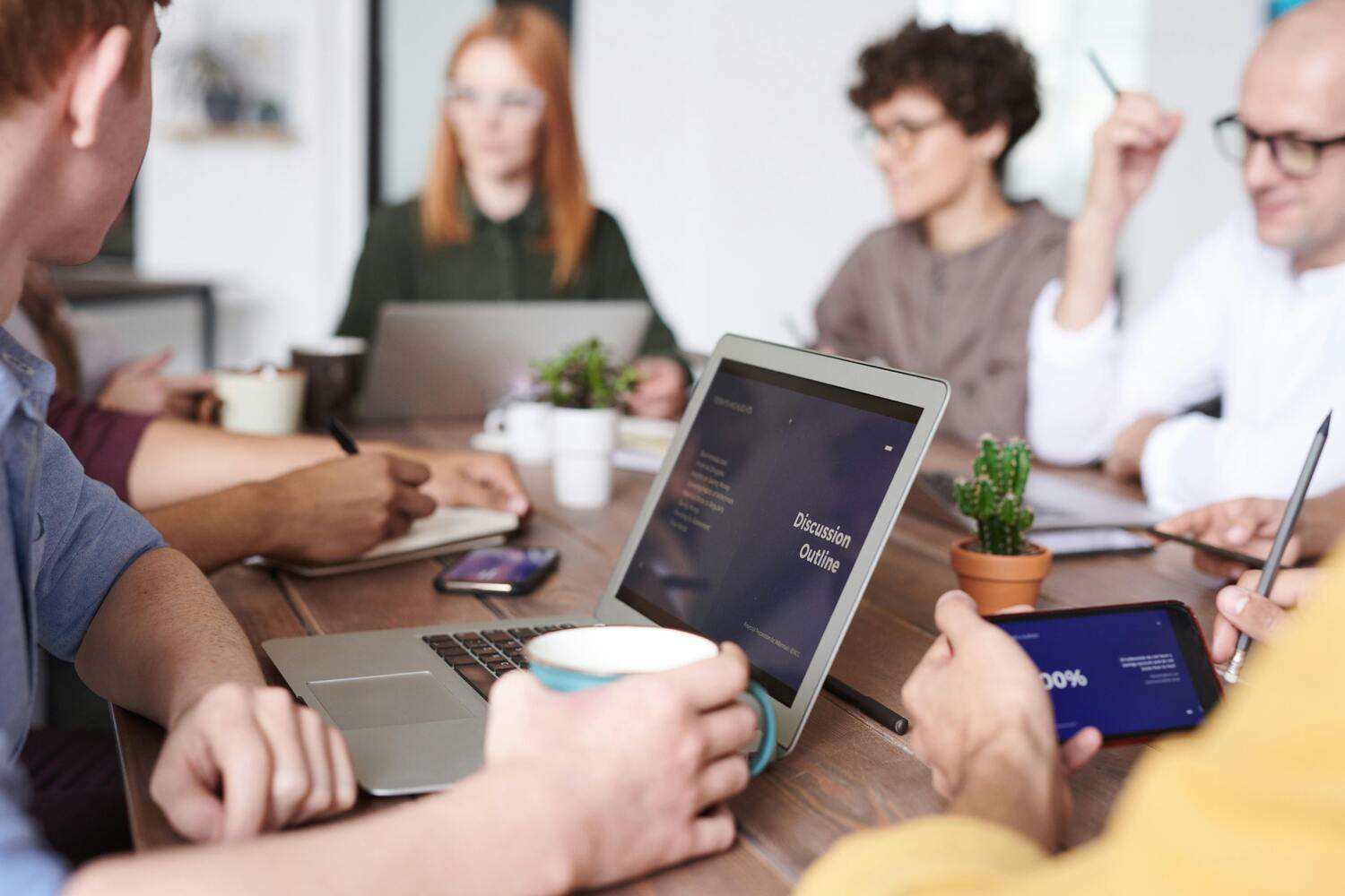 Réunion d'entreprise avec un focus sur le matériel informatique disposé sur la table ; tablettes, smartphones, laptops. Les personnes sont floues.
