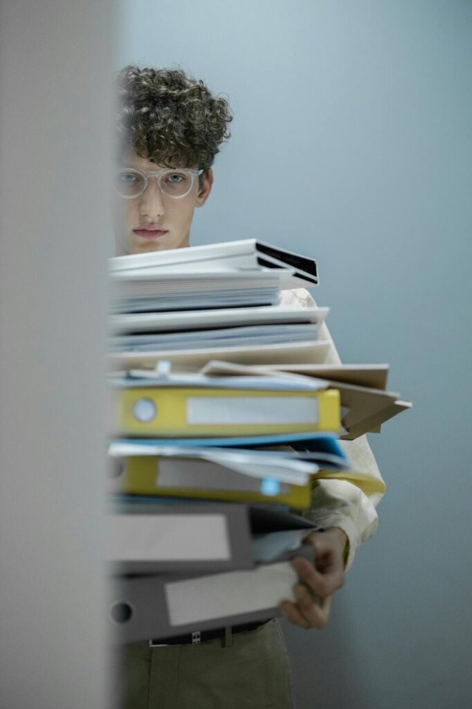 Un jeune administratif est debout les bars chargés d'une haute pile de classeurs et de dossiers