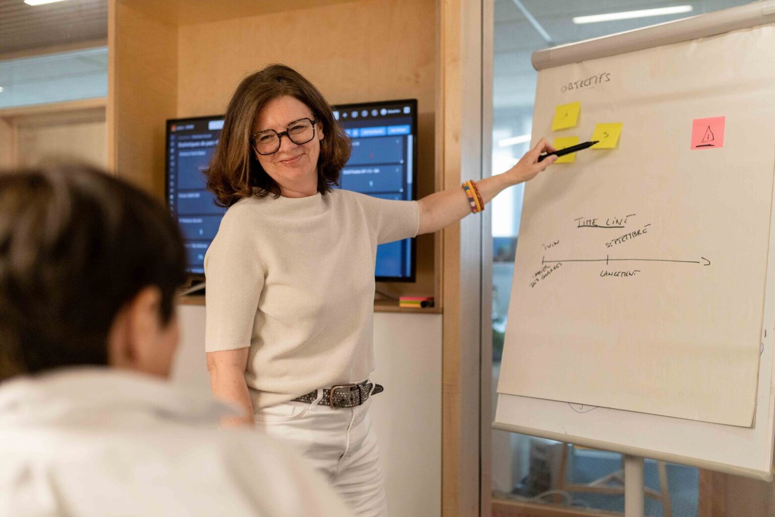 Une personne de l'équipe Kubs est debout devant le paperboard et pointe des post-it et objectifs. La cliente est tournée vers le paperboard, assise, elle écoute attentivement les explications.
