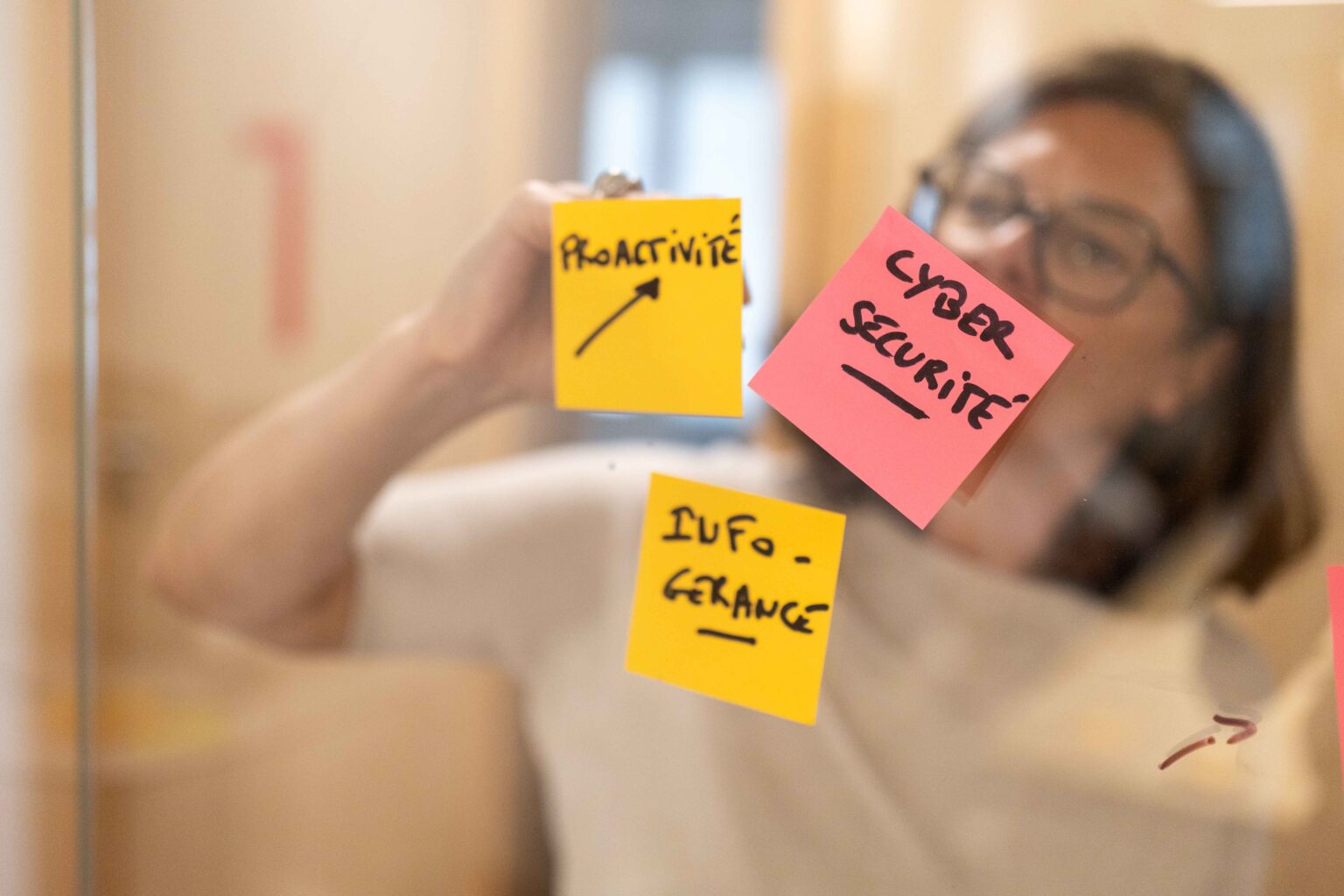 En arrière plan une personne floutée écrit sur une surface vitrée, sur laquelle des post-it sont collés en premier plan. On peut lire sur les post-it les inscriptions suivantes : Cybersécurité, infogérance et Proactivité