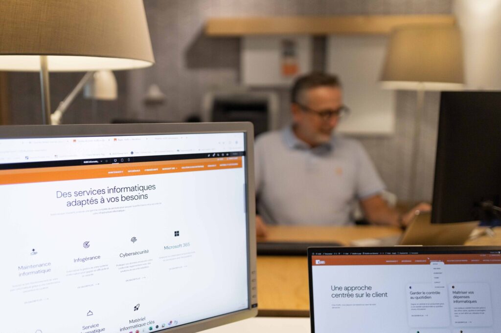 Focus sur 2 écrans d'ordinateur chez Kubs. Une personne de l'équipe en second plan floutée au fond. Sur le premier écrans sont présentés les services informatiques Kubs et, sur le second, l'approche de Kubs centrée sur les spécificités de chaque client