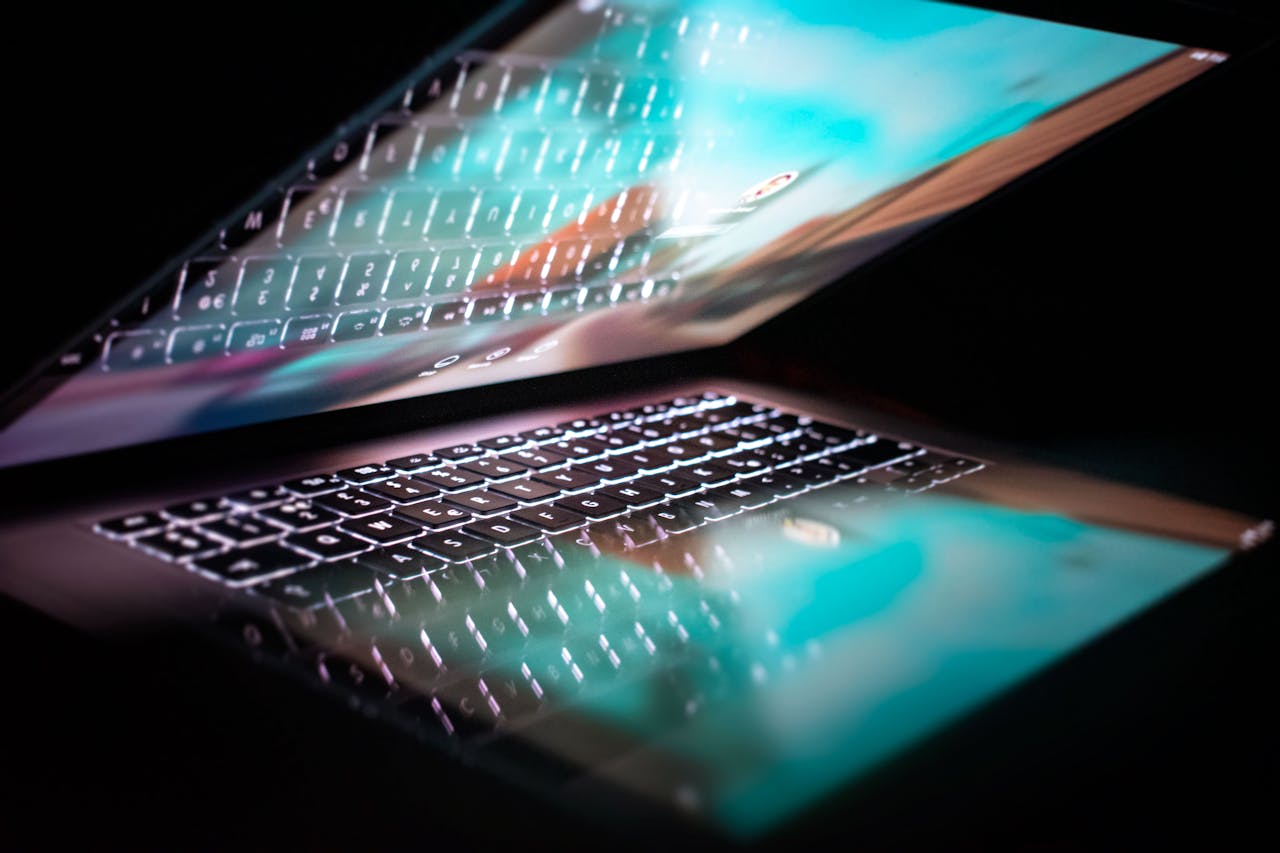 Zoom sur une PC portable entrouvert. L'écran est allumé et le clavier rétroéclairé se reflète dans l'écran. L'arrière plan est noir